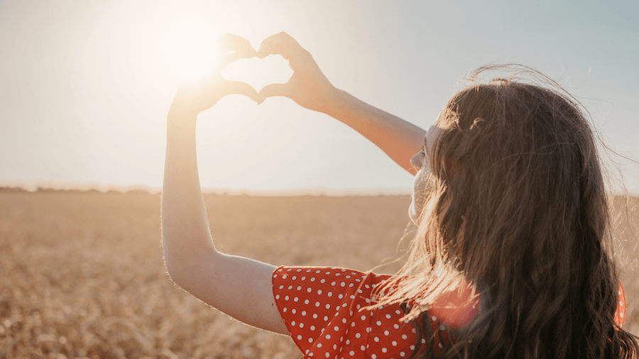 Image showing a black haired girl holding a heart shape at the sun in an open field