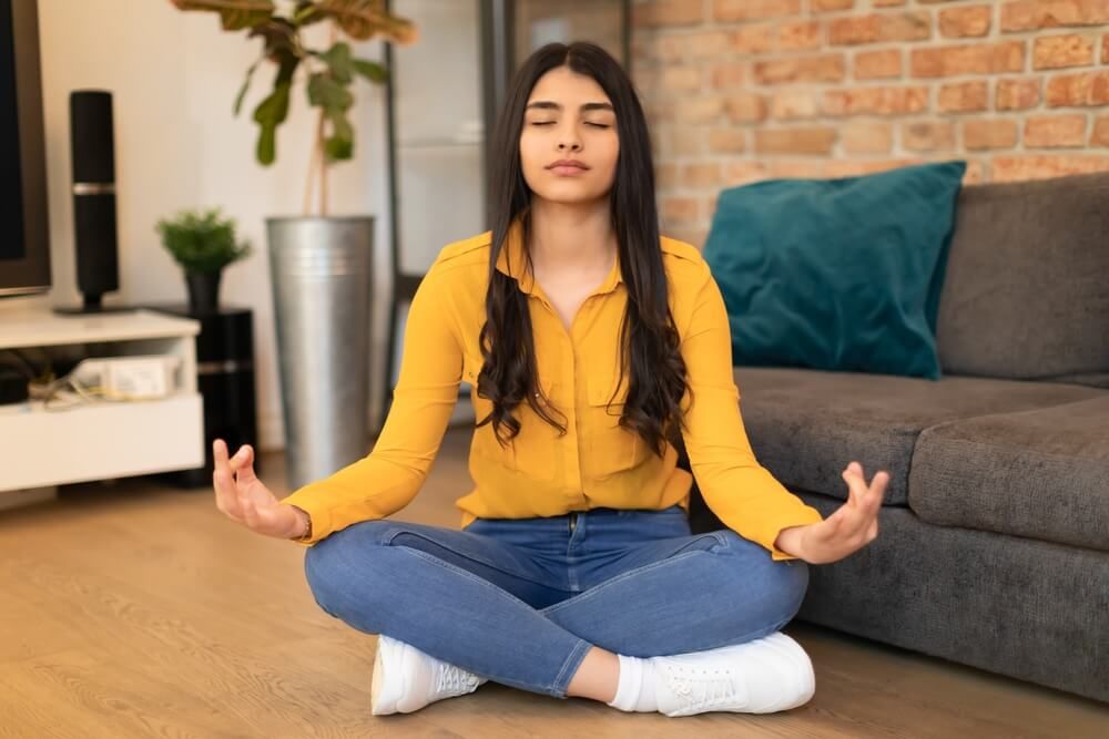 Calm Spanish lady meditating with closed eyes, sitting on floor at home in living room for managing slow COMT stress