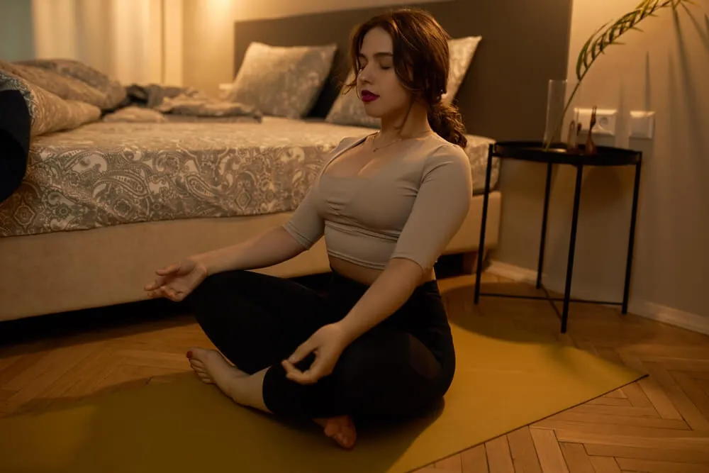 A person doing a relaxing breathing technique on the floor in their bedroom to fall asleep faster. They are sitting in a lotus posture with closed eyes, dressed in black leggings and a beige sports top.