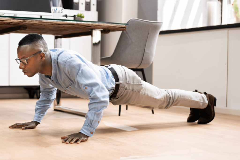 A man exercise snacking at his office by doing a quick workout near his desk. He is seen in formals doing push ups on the floor next to his desk.