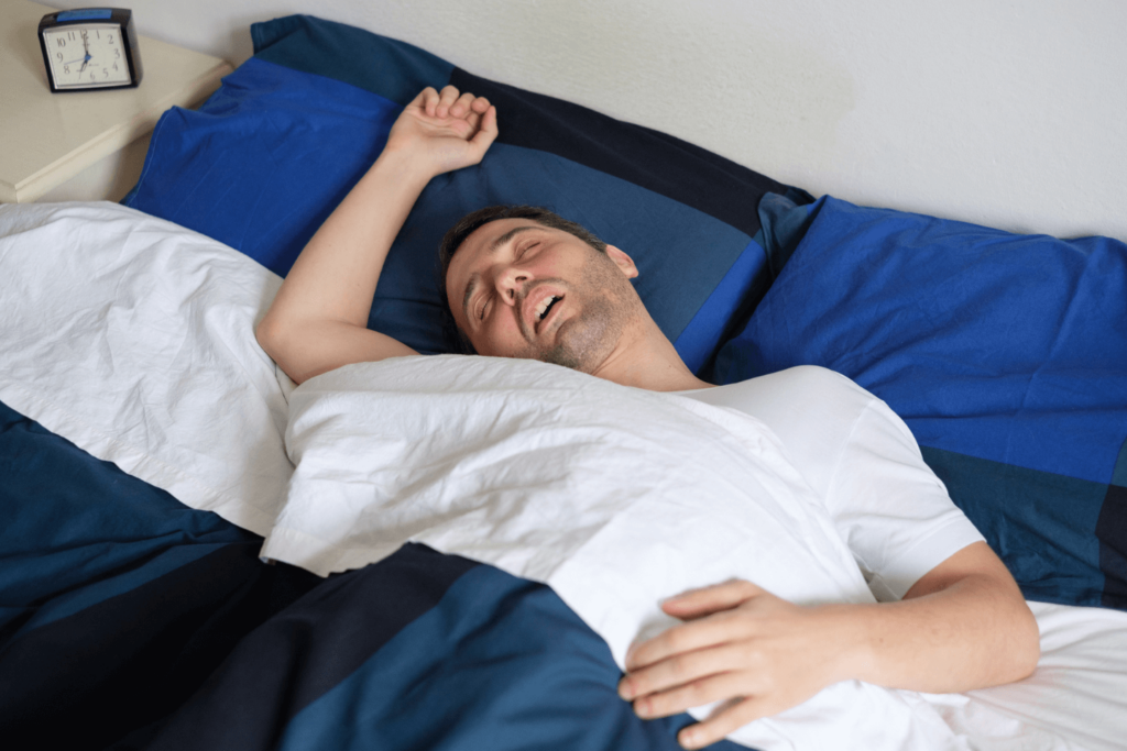 Can sleep apnea lead to brain damage? A person sleeping on their bed with an arm up and mouth open, indicating that they are snoring.