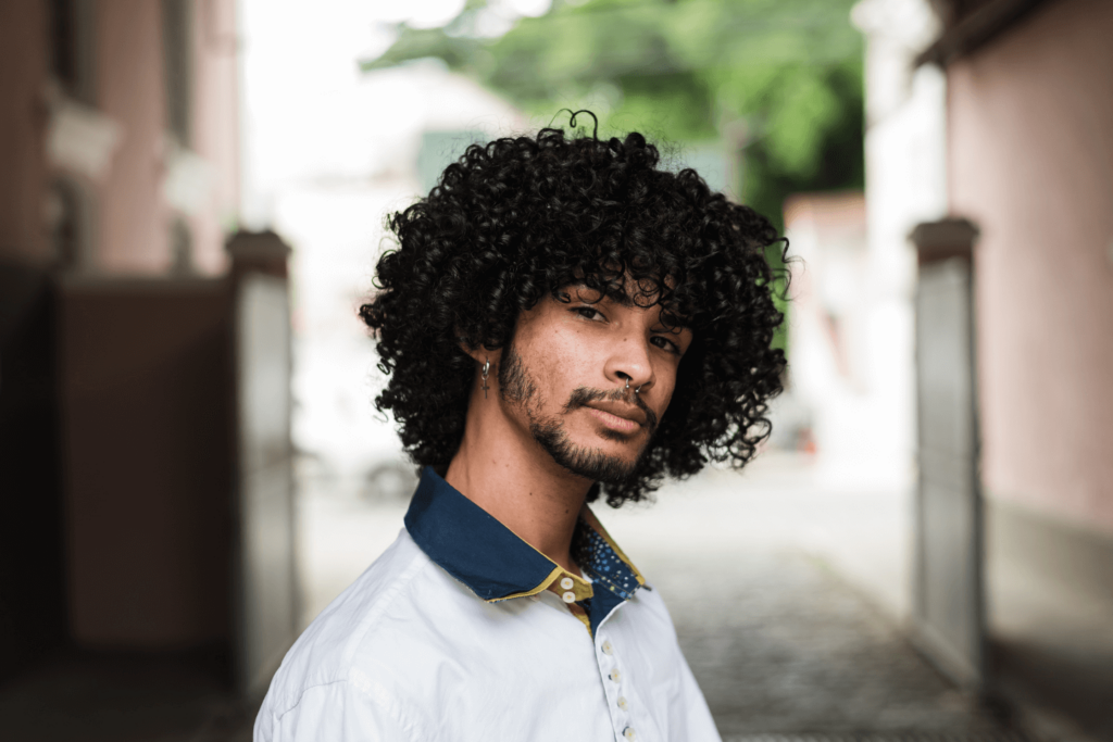 A curly haired male in a white polo T-shirt with a dark blue collar standing in what appears to be a street.