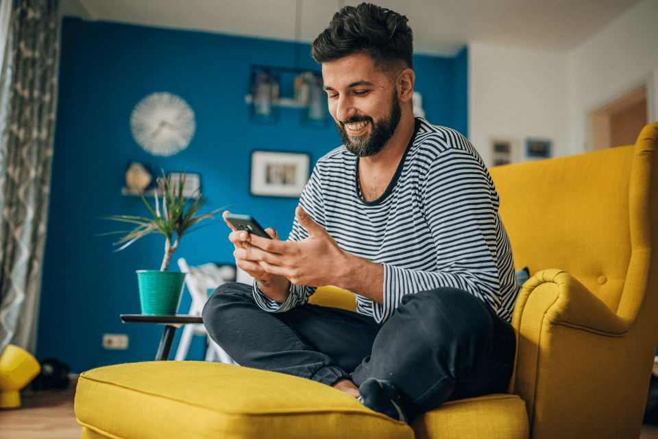 A young man look at his smartphone and smiling. He's sitting on his bright yellow couch in the living room. Could excessive use of smart phones cause infertility?