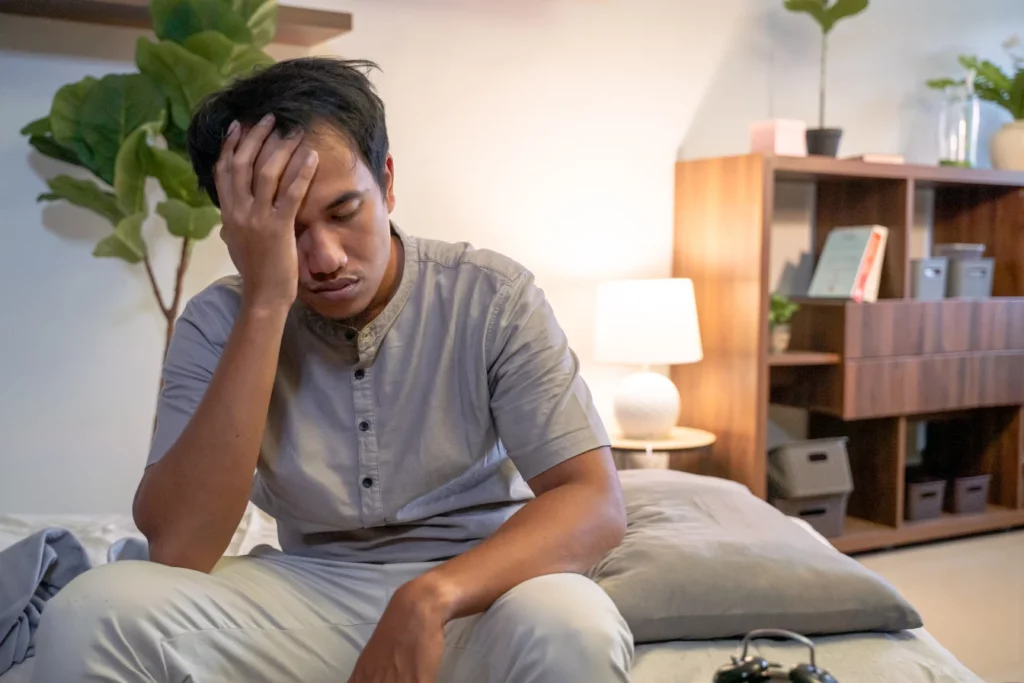 A man in his bedroom sitting on his bed with his hand on the head indicating that he's sleep deprived. Lack of sleep can cause many gastrointestinal issues, including nausea.