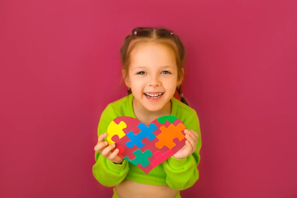 A girl child holding a heart made of legos. She is happily smiling and is against a red background.