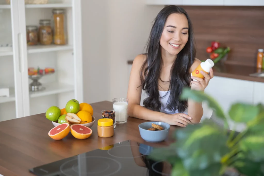 A woman leaning on her kitchen countertop holding a bottle of supplements with her left hand. Her right hand is resting on the counter top. There are some citrus fruits on the table.