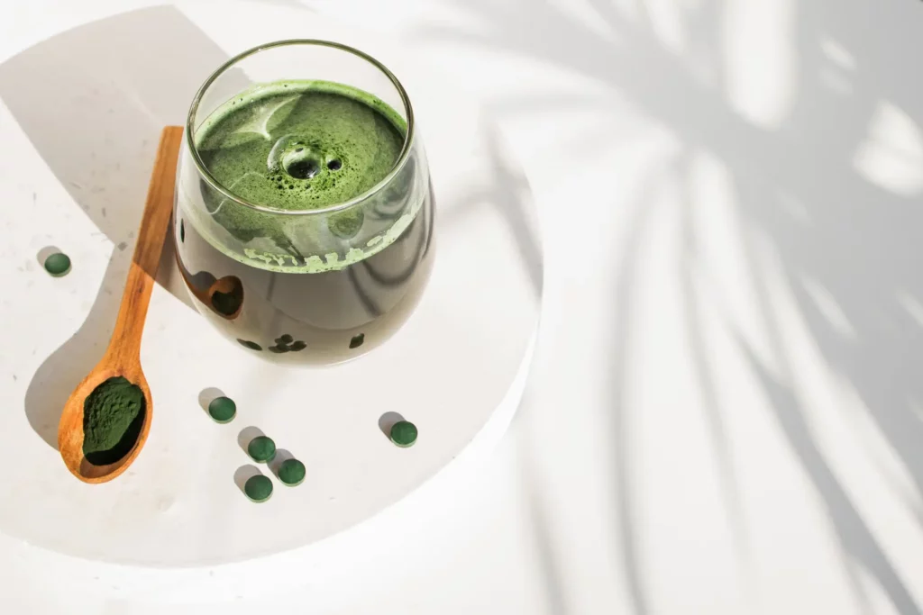 Spirulina powder mixed in a glass of water placed on a plate along with a wooden teaspoon and some spirulina tablets