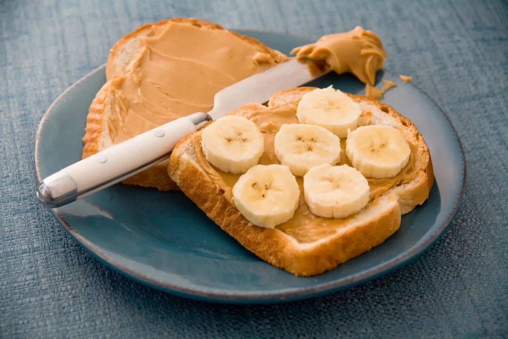 2 slices of peanut butter toast. On one slice, there are some cut pieces of banana. Along with a butter knife the toasts are on a plate.